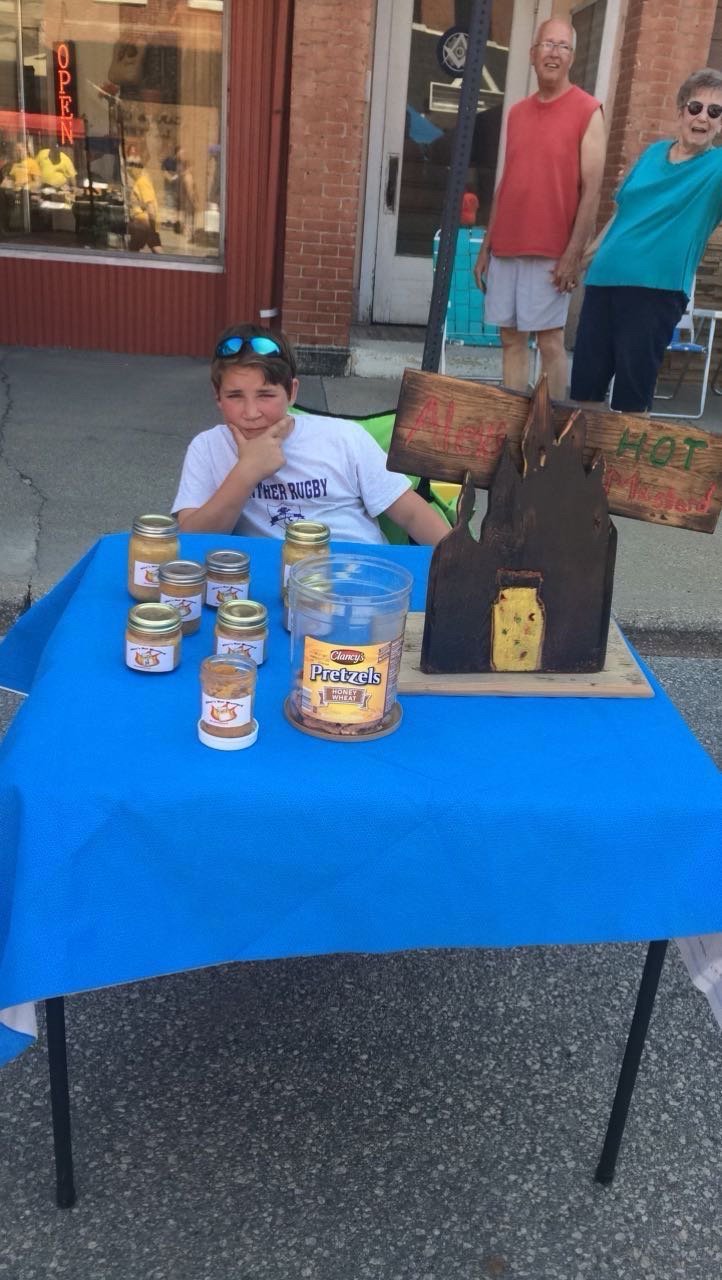 A young Alex sitting behind a blue table of mustard jars with a hand-lettered Alex's Hot Mustard sign and a tub of pretzels