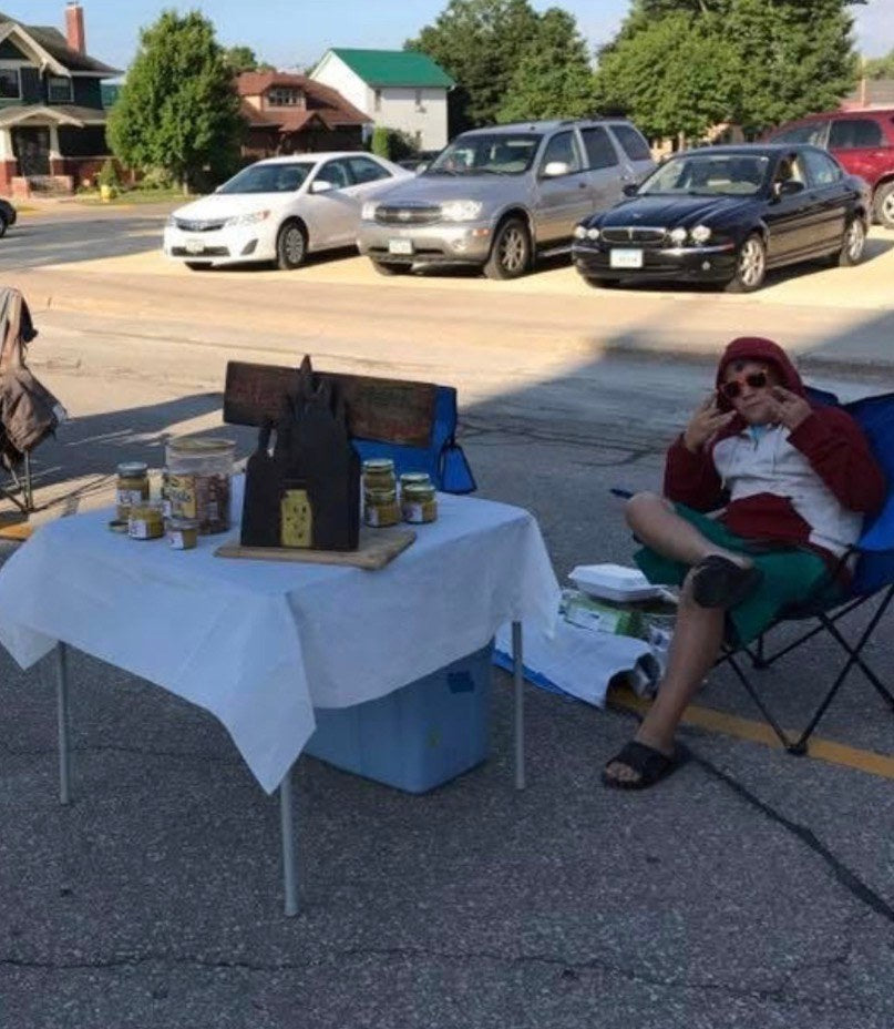 Alex in a folding chair beside a table of jars and a hand-lettered Alex's Hot Mustard sign at an early market stand