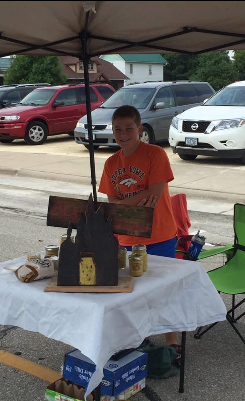 Alex under a market tent in an orange Denver Broncos shirt next to his hand-made sign